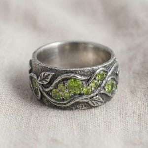 Könnte beinhalten: Ein silberner Ring mit einem strukturierten Band, das ein Blatt- und Weinreben-Design aufweist. Der Ring ist mit mehreren runden, hellgrünen Edelsteinen verziert. Der Ring ist vor einem neutralen Hintergrund platziert.
