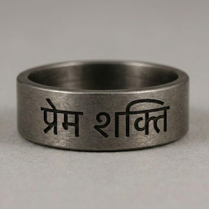 Könnte beinhalten: Ein dunkelgrauer Metallring mit gebürsteter Oberfläche und einer schwarzen Gravur in Devanagari-Schrift, die Prem Shakti lautet. Das Band hat ein flaches Profil und eine gleichmäßige Breite über den gesamten Umfang mit einer metallischen Textur.