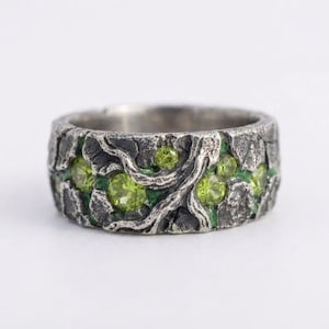 May include: A wide, textured silver ring with a dark, bark-like pattern. The ring features bright green gemstones set within the design, with a touch of green enamel. The ring's design evokes a natural, organic theme.