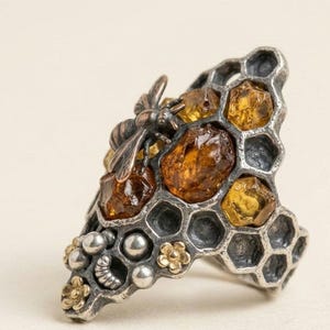 Puede incluir: Un anillo de plata con un patrón de panal y piedras incrustadas de color ámbar. Una abeja esculpida se asienta sobre las celdas hexagonales, acompañada de pequeñas esferas plateadas y diminutos detalles de flores en metal oxidado.
