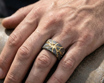 May include: A textured, wide band ring with a dark gray base and gold-colored accents. The ring's design features a starburst pattern. The ring is shown on a person's hand.