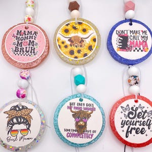 May include: A collection of six round car air fresheners with various designs and text. The designs include sunflowers, a Highland cow, and phrases like "Mama Mommy Mom Bruh" and "Set yourself free." Each has a beaded string.