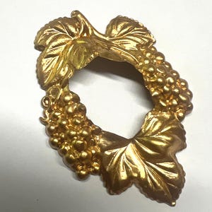 May include: A gold-colored, grape-themed decorative item. The design features a cluster of grapes and leaves, forming a circular shape. The item is likely made of metal and has a shiny finish, suggesting it could be a brooch or decorative element.