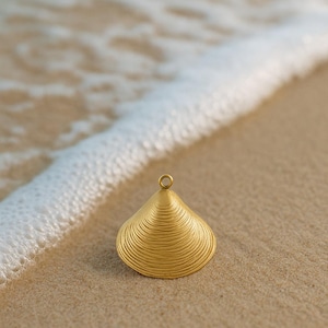 May include: A gold-colored seashell pendant with a textured surface, resting on a sandy beach. The pendant has a small loop at the top. Gentle waves with white foam are in the background.