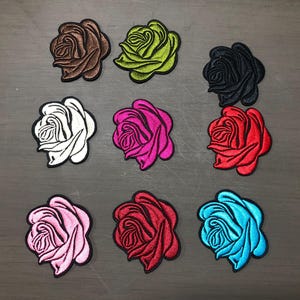 Puede incluir: Nueve parches de rosas bordadas en varios colores, incluyendo marrón, verde, negro, blanco, rosa, rojo y azul. Cada parche presenta un diseño floral detallado con un borde negro, adecuado para la decoración de telas.