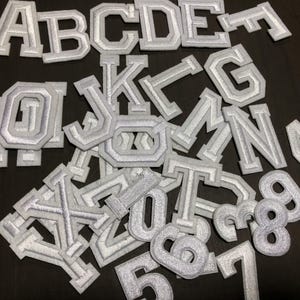 Puede incluir: Una colección de letras y números de tela bordada blanca. Las letras incluyen A, B, C, D, E, G, J, K, L, M, N, O, T y los números 4, 5, 6, 7, 8 y 9. Los parches son de estilo universitario.