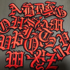 Może przedstawiać: Kolekcja czerwonych, haftowanych naszywek z gotyckim alfabetem z czarnymi obwódkami. Litery są w odważnej, stylizowanej czcionce i są rozrzucone na szarej powierzchni. Naszywki prawdopodobnie służą do personalizacji odzieży lub akcesoriów.