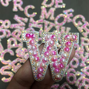 Puede incluir: Una letra "W" blanca adornada con perlas rosas y blancas, rodeada de pedrería iridiscente. Otras letras son visibles en el fondo. Probablemente para manualidades o adornos.
