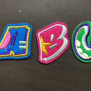 Op de afbeelding: Drie geborduurde stoffen patches in de vorm van de letters A, B en C. De letter A is blauw met roze en paarse accenten. De letter B is roze met een gele ster. De letter C is groen met witte stippen en paarse accenten.