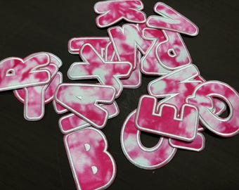 May include: A collection of pink and white tie-dye fabric letters with a white border. The letters are in various fonts and sizes, scattered on a dark surface. The letters appear to be patches or appliques.