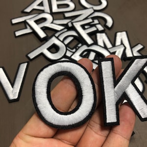 Pode incluir: Letras brancas bordadas com bordas pretas, incluindo as letras V, O e K, são exibidas em um fundo neutro. Outras letras estão espalhadas no fundo. As letras são provavelmente patches ou apliques.