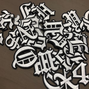 Op de afbeelding: Een verzameling witte geborduurde patches met gotische letters en zwarte randen. De patches, in verschillende stijlen, zijn verspreid over een oppervlak, met de verschillende letterontwerpen.
