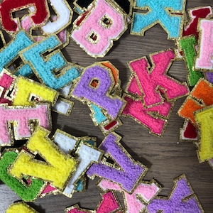 Könnte beinhalten: Eine Sammlung bunter, gestickter Stoffbuchstaben in verschiedenen Farben, darunter Rosa, Gelb, Blau und Lila. Jeder Buchstabe ist mit goldfarbenem Zierrat umrandet. Die Buchstaben sind auf einer Holzoberfläche verstreut.