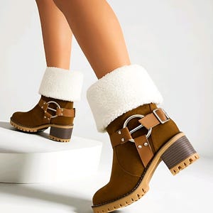 May include: Brown suede ankle boots with a cream-colored faux fur cuff. The boots feature a chunky heel, a light brown sole, and a decorative buckle and strap detail. The boots are designed for cold weather.