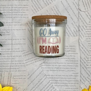 Op de afbeelding: Een glazen potkaars met een houten deksel. De kaars heeft een wit interieur en een label met de tekst "GO Away (I'M) READING". De kaars is waarschijnlijk geparfumeerd en ontworpen voor ontspanning.