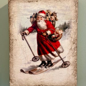 May include: A rectangular wall plaque featuring a vintage-style illustration of Santa Claus skiing on snow. Santa wears a red coat with white fur trim, carries a sack of toys and a basket of apples, and uses wooden ski poles on a cracked, aged background.