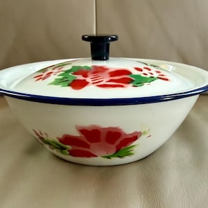 May include: White enamelware bowl with a lid, featuring a floral design in red and green. The bowl has a blue rim and a black handle on the lid. A classic kitchenware item.