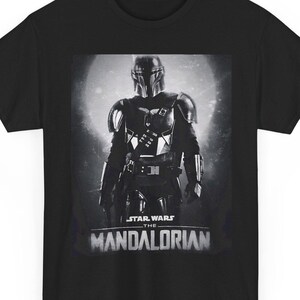 Può includere: T-shirt nera con un'immagine in scala di grigi di The Mandalorian di Star Wars. L'immagine mostra il personaggio con l'armatura completa su uno sfondo illuminato dalla luna. Le parole "STAR WARS" e "THE MANDALORIAN" sono stampate sotto.
