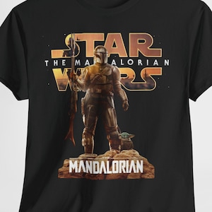 Può includere: T-shirt nera con grafica della serie Star Wars "The Mandalorian". Il design include il titolo dello show, un personaggio con un'arma e una piccola creatura verde. Il design utilizza colori oro, marrone e nero.