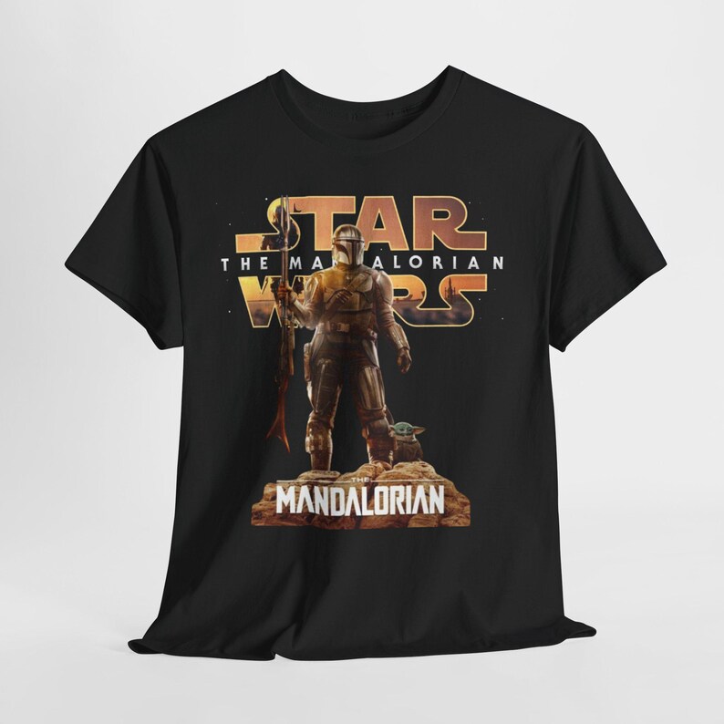 Può includere: T-shirt nera con grafica della serie Star Wars "The Mandalorian". Il design include il titolo dello show, un personaggio con un'arma e una piccola creatura verde. Il design utilizza colori oro, marrone e nero.