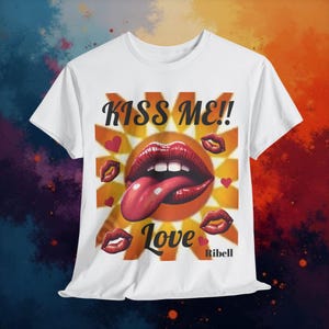 Può includere: T-shirt bianca con un design grafico. Il design include il testo "KISS ME!!" sopra un paio di labbra rosse con una lunga lingua. La parola "Love" è sotto le labbra, con piccoli cuori rossi e grafica di labbra aggiuntiva. Il marchio "Ribell" è in basso.