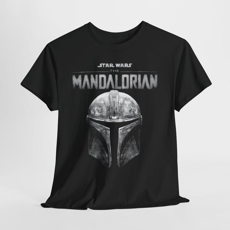 Può includere: T-shirt nera con la scritta "STAR WARS THE MANDALORIAN" e un'illustrazione dettagliata del casco del Mandaloriano. Il casco è grigio con un aspetto usurato. La maglietta è realizzata in materiale morbido.