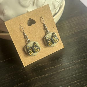 Könnte beinhalten: Handgefertigte Ohrringe mit einem Schmetterlingsdesign. Jeder Ohrring hat eine sechseckige Form mit hellgrünem Hintergrund und einem detaillierten Schmetterling in Gelb, Schwarz und Orange. Die Ohrringe sind an silberfarbenen Haken befestigt und werden auf einer braunen Karte präsentiert.