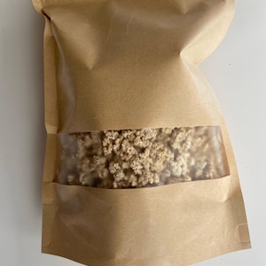 May include: A brown paper bag with a clear window displaying dried, beige-colored flowers. The bag is sealed at the top with a zipper closure. The bag is sitting on a white surface.
