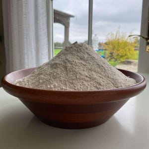 Könnte beinhalten: Eine Holzschüssel gefüllt mit einer großen Menge hellbraunem Mehl. Die Schüssel hat eine satte, dunkelbraune Farbe und steht auf einer weißen Oberfläche. Der Hintergrund zeigt ein Fenster mit Blick auf einen bewölkten Himmel und Bäume.