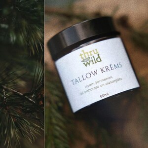 Op de afbeelding: Een witte pot met een zwarte deksel en een groen-wit etiket met de tekst "thru wild TALLOW KRĒMS visam kērmenim, lai pabarotu un aizsargātu 60ml".