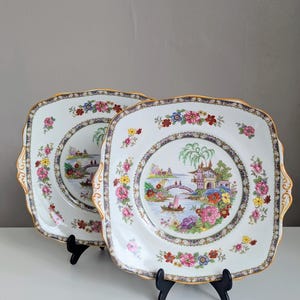 May include: Two square, white ceramic plates with a gold trim and a colorful floral and landscape design. The plates feature a central scene with a bridge, boats, and buildings, surrounded by a border of flowers.