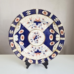 Puede incluir: Un plato de cerámica redondo con una paleta de colores azul oscuro y crema con intrincados patrones florales. El diseño incluye un motivo circular central rodeado por cuatro paneles alternos de azul y crema, acentuados con detalles en naranja y dorado.