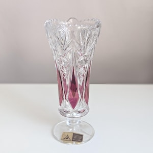 Peut inclure: Un vase en cristal transparent avec un intérieur rouge rubis. Le vase présente un motif détaillé en cristal taillé et un bord festonné. Il repose sur une base claire et circulaire. Une petite étiquette est apposée sur la base.