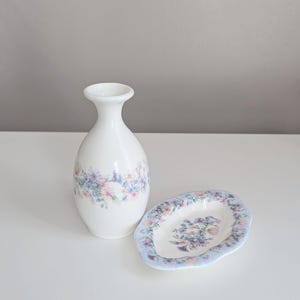 May include: A white ceramic vase and a small oval dish. Both items feature a floral pattern in pastel pink, blue, and purple. The vase has a slender neck and a rounded body. The dish has a scalloped edge.
