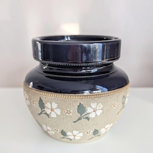 Può includere: Un vaso di ceramica blu scuro con un'ampia apertura e un corpo arrotondato. La parte inferiore presenta una fascia beige ornata da motivi floreali bianchi e foglie verdi. Il vaso ha una finitura lucida e un bordo decorativo.