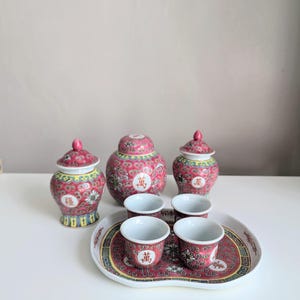Peut inclure: Un service à thé chinois vintage avec un motif rouge et floral. L'ensemble comprend trois pots à couvercle, quatre petites tasses et un plateau de service. Les tasses et les pots ont des caractères chinois. L'ensemble est sur une surface blanche.