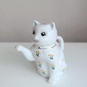 Puede incluir: Una tetera blanca de cerámica con forma de gato y una pata levantada. La tetera está decorada con motivos florales amarillos y un collar dorado. El gato tiene ojos negros y nariz rosa.