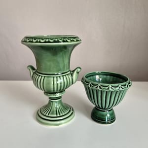 Op de afbeelding: Twee saliegroene keramische vaten met een glanzende afwerking. Het hogere stuk is een urn op een voet met twee kleine handvatten aan de zijkanten en een uitlopende rand. Het kortere stuk is een kom op een voet met een geschulpte rand.