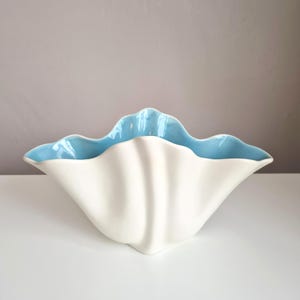 Puede incluir: Un cuenco de cerámica con forma de concha marina, con un exterior blanco liso y un interior azul claro brillante. El cuenco presenta un borde ondulado y crestas verticales en los laterales, creando una forma abierta con una silueta curva.
