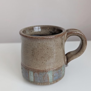 Könnte beinhalten: Eine handgefertigte Keramiktasse mit brauner, gesprenkelter Glasur. Die Tasse hat einen abgerundeten Henkel und ein blau-grünes Band am Boden. Die Tasse ist etwa 10 cm hoch.