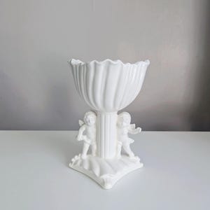 Könnte beinhalten: Eine weiße Keramik-Sockelschale mit geriffeltem Design und gewelltem Rand. Zwei Cherubfiguren stützen die Basis der Schale. Die Schale steht auf einer weißen Oberfläche vor einem grauen Hintergrund.