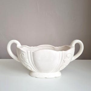 Może przedstawiać: Biała ceramiczna miska z dwoma zakrzywionymi uchwytami po przeciwnych stronach. Miska posiada falisty brzeg oraz tłoczone wzory w kształcie zwojów na zewnętrznych ściankach. Spoczywa na małej, okrągłej podstawie z fakturowanym wykończeniem.