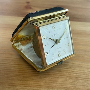 Op de afbeelding: Een vintage Europa reiswekker. De klok heeft een goudkleurige metalen behuizing met een witte wijzerplaat, zwarte cijfers en gouden wijzers. De klok is open en onthult de wijzerplaat en een kleine, goudkleurige bel. De behuizing is gevoerd met een bloemenstof.