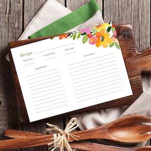 May include: A white recipe card with floral watercolor accents, featuring orange, pink, and yellow flowers. The card includes sections for prep time, cook time, ingredients, servings, and directions. It rests on a wooden cutting board with wooden spoons and a linen napkin.