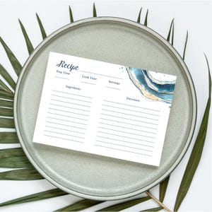 May include: A white recipe card with the word "Recipe" in cursive script at the top. The card has lines for ingredients and directions, with a blue and gold watercolor design in the corner. The card is on a gray plate with green leaves.