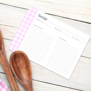 Puede incluir: Una tarjeta de recetas con un borde rosa y blanco a cuadros. La tarjeta tiene líneas para ingredientes, tiempo de cocción, instrucciones y porciones. La palabra "Receta" está impresa en la parte superior. Dos cucharas de madera están en primer plano.