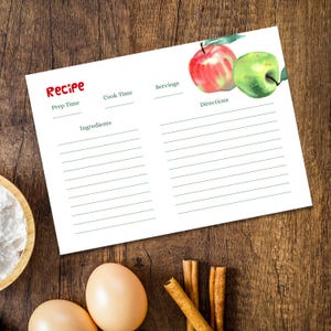 May include: A white recipe card with the word "Recipe" in red, featuring a watercolor illustration of a red and green apple. The card has sections for prep time, cook time, servings, ingredients, and directions. Cinnamon sticks, eggs, and flour are also visible.