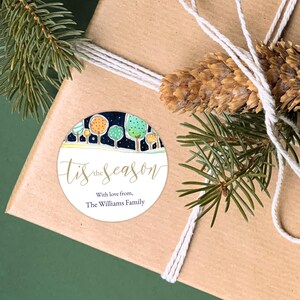 May include: A Christmas gift wrapped in brown paper, adorned with a round sticker saying "Tis the Season". The sticker shows a watercolour illustration of trees under a starry night. The gift is decorated with twine, pine needles, and a pine cone.