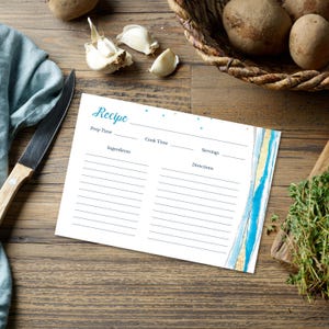 May include: A white recipe card with the word "Recipe" in teal script. The card has lines for ingredients, prep time, cook time, servings, and directions. It is decorated with blue and gold watercolor stripes. The card is on a wooden surface with potatoes, garlic, and a knife.