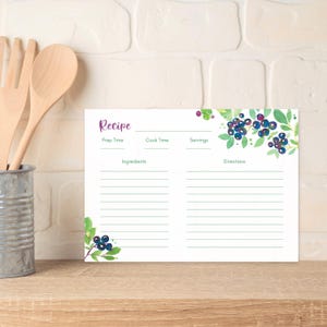 May include: A white recipe card with a watercolor design of blueberries and green leaves. The card has sections for prep time, cook time, servings, ingredients, and directions. Wooden utensils are in a metal container to the left.